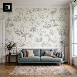 Puede incluir: Un sofá azul claro se sitúa frente a una pared con papel pintado blanco roto que presenta un patrón chinoiserie en azul y verde de pagodas, puentes y sauces. El sofá incluye dos cojines beige lisos y un cojín marrón texturizado.