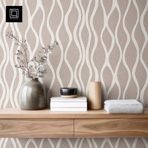 May include: A neutral-toned interior scene featuring a wooden shelf with decorative items. A vase with branches, a stack of books, a small bowl, a wooden cylinder, and a folded blanket are arranged on the shelf. The background is a textured wallpaper with a wavy pattern.