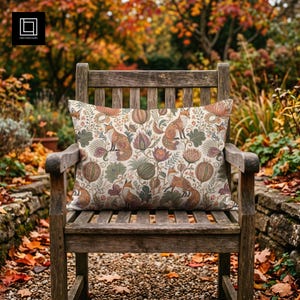 May include: A rectangular throw pillow featuring a repeating pattern of foxes, stylised flowers, and foliage in muted tones of orange, green, and brown on a cream background, resting on a wooden chair in an outdoor setting.