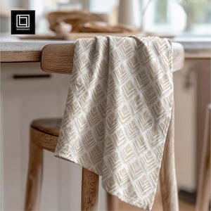 May include: A beige and white patterned kitchen towel draped over a wooden chair. The towel has a repeating geometric design. The chair is made of light-coloured wood, and the background is a blurred kitchen scene.