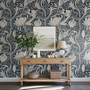 May include: A wooden console table with three drawers, a framed picture, a lamp, a vase with white flowers, a woven basket, and a round cushion. The table is in front of a wall with a dark blue and white floral wallpaper.