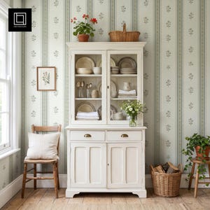 May include: A white wooden hutch with glass-front upper cabinets and two lower drawers and doors. The shelves contain stacked plates, bowls, glassware, and a small vase of flowers. A wicker basket sits on top and another on the floor beside it.