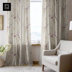May include: Sheer, floral-patterned curtains in cream, beige, and purple hang in front of a window. The curtains feature a delicate botanical design, with a white armchair in the foreground. The room is well-lit, creating a soft and inviting atmosphere.