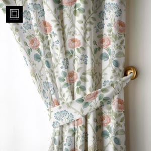 Può includere: Tenda con motivo floreale e fermatenda. La tenda bianca presenta un motivo ripetuto di rose rosa, fiori blu e foglie verdi. È visibile un fermatenda dorato.