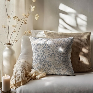 May include: A white sofa with two cushions. The sofa has a throw draped over the arm. One cushion is a light blue and cream patterned cushion with a damask design. The other cushion is a brown woven cushion. The sofa is in a room with a white vase with dried branches.