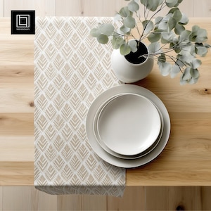 May include: A table setting featuring a cream-colored table runner with a geometric leaf pattern. Two white plates are stacked on top of each other. A white vase holds eucalyptus branches. The table is made of light-colored wood.