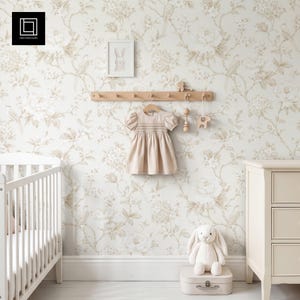 May include: A beige smocked dress hangs on a wooden wall rack against floral wallpaper. A white framed rabbit print, a wooden rattle, a wooden animal toy, a white plush rabbit, and a small suitcase are displayed in a nursery setting.