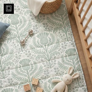 Può includere: Un tappeto gioco per bambini con un motivo floreale di cardo in verde tenue e bianco. Un sonaglio d'argento, blocchi di lettere di legno e un coniglietto di peluche sono sul tappeto. Un cestino di vimini e una culla sono sullo sfondo.