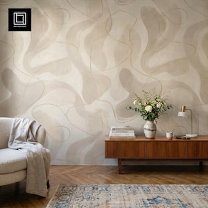 May include: A room interior featuring wallpaper with a beige and cream abstract fluid pattern and thin gold lines. A wooden sideboard holds a white vase with flowers and books. A textured armchair with a throw blanket sits on a patterned rug.