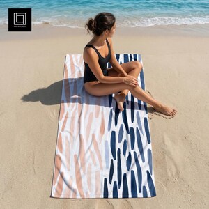 May include: A beach towel with a white background and a pattern of vertical brushstrokes in shades of peach, light blue, and navy blue. The towel is rectangular and is on a sandy beach near the ocean.