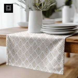 May include: A beige and white patterned table runner with a geometric design. The runner is placed on a wooden table, with white plates and a vase of greenery in the background. The table runner is made of fabric.