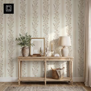 May include: A light wood console table with a lower shelf, topped with a ceramic vase containing greenery, a framed abstract print, a small bust, a candle, and a lamp. A woven basket sits on the lower shelf against floral striped wallpaper.