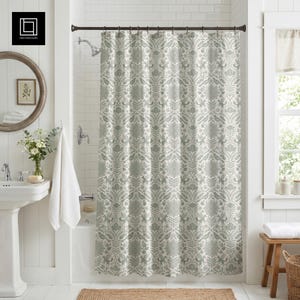Peut inclure: Rideau de douche à motifs floraux gris et blancs. Il est suspendu à une tringle marron foncé dans une salle de bain blanche. On aperçoit un lavabo sur pied, un miroir rond et une fenêtre avec un banc en bois.