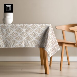 May include: A rectangular tablecloth with a geometric pattern of beige and white diamonds covers a wooden table. A wooden chair is positioned to the right. The tablecloth is made of a soft, draping fabric.