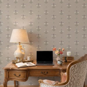 May include: A wooden desk with a beige lamp, a laptop, an open notebook with a pen, a small vase with flowers, a candle, and a stack of books. The desk is positioned against a wall covered in wallpaper featuring a repeating floral pattern.