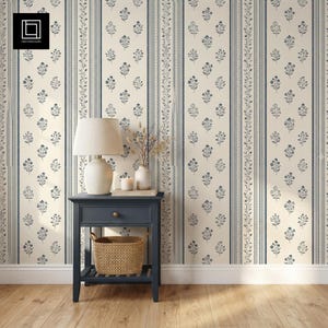Può includere: Un tavolino in legno blu scuro con un cassetto e un ripiano inferiore che ospita un cesto intrecciato. Sopra si trovano una lampada da tavolo color crema, una piccola candela bianca e un vaso con steli secchi su carta da parati a righe.