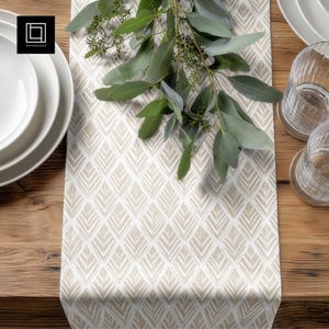 May include: A beige and white patterned table runner with a geometric design, featuring a repeating leaf-like pattern. The runner is placed on a wooden table, with white plates and clear glasses visible. Green eucalyptus branches are draped across the runner.