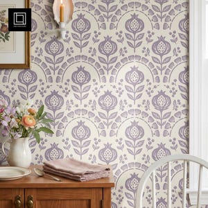 May include: A room with a cream-colored wall featuring a repeating pattern of stylized purple pomegranates and floral elements. A wooden cabinet holds a white pitcher of flowers, a plate, and a folded pink napkin. A lit candle sits on the wall.