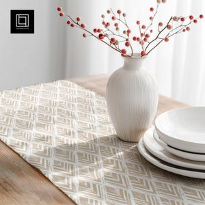 Może przedstawiać: Beżowo-biały bieżnik w geometryczny wzór na drewnianym stole. Biały wazon z czerwonymi jagodami stoi obok stosu białych talerzy. Obraz jest dobrze oświetlony, ukazując tekstury i wzory.