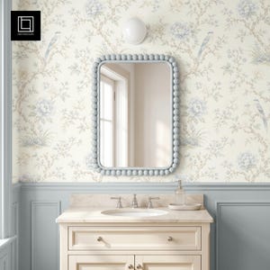 May include: A rectangular wall mirror with a light blue beaded frame, mounted above a cream-colored bathroom vanity with a white sink, chrome faucet, and a light blue wainscoted wall featuring floral and bird wallpaper in blue and cream tones.