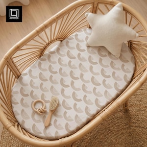 Puede incluir: Una cuna de mimbre con una sábana bajera estampada, una almohada en forma de estrella, un cepillo para bebés de madera y un mordedor de madera. La sábana presenta un patrón repetido de semicírculos beige sobre un fondo crema. La cuna está sobre una alfombra tejida.