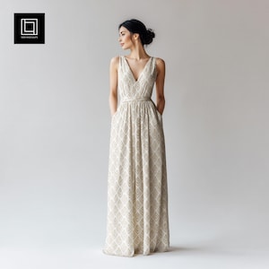 May include: A long, sleeveless dress with a V-neck and a geometric pattern in beige and white. The dress has a fitted waist and pockets. The model is wearing the dress and has her hands in the pockets.