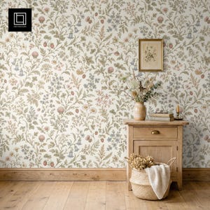 Pode incluir: Um quarto com papel de parede floral com fundo creme e um padrão de flores e folhas em tons de rosa, azul e verde. Uma mesa de cabeceira de madeira com um quadro emoldurado, um vaso e uma cesta está em frente ao papel de parede.