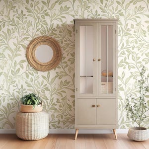 May include: A light gray cabinet with glass doors and wooden knobs. The cabinet is standing on a wooden floor in front of a wall with a green floral wallpaper pattern.