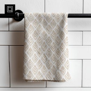 Op de afbeelding: Een beige en wit patroon handdoek hangt aan een zwarte stang. De handdoek heeft een herhalend geometrisch ontwerp van bladachtige vormen. De achtergrond is witte tegels.
