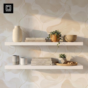 May include: Two white floating shelves against a beige abstract wallpaper. The top shelf holds a tall beige vase, two books titled Modern Forms and Earth Tones, a potted green plant in a terracotta pot, and a beige bowl. The bottom shelf holds two gray cups, three books titled Sculptural Space, Ceramic Art, and Living Well, a small succulent in a geometric pot, and a round woven tray with small wooden spheres.