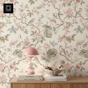 Könnte beinhalten: Tapete mit einem sich wiederholenden Muster aus weißen Kranichen, kleinen Vögeln, rosa Blüten und grünen Blättern auf cremefarbenem Grund. Eine rosa kuppelförmige Lampe steht auf zwei Büchern mit der Aufschrift Interiors und Design Curation.