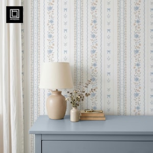 Könnte beinhalten: Eine beige Lampe mit cremefarbenem Lampenschirm steht auf einer hellblauen Kommode. Der Hintergrund ist eine Tapete mit vertikalen Streifen mit Blumen- und Schleifenmustern in Blau, Beige und Weiß. Eine kleine Vase und Bücher stehen auf der Kommode.