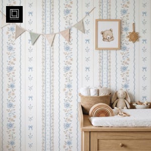Pode incluir: Um interior de berçário com papel de parede floral em tons de azul e bege. Uma mesa de mudança de madeira com uma cesta de artigos para bebês, um coelho de pelúcia e um arco-íris de madeira. Uma impressão de urso emoldurada e uma guirlanda de bandeiras decoram a parede.