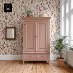 Könnte beinhalten: Ein hellrosa Holzschrank mit zwei Türen und zwei Schubladen. Der Schrank befindet sich in einem Raum mit floraler Tapete und Holzboden. Eine Topfpflanze und ein Spiegel sind ebenfalls sichtbar.