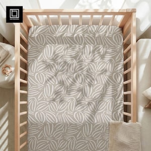 May include: A fitted crib sheet with a neutral beige background and a repeating pattern of white, abstract leaf shapes. The sheet is inside a light-colored wooden crib. The overall aesthetic is minimalist and modern.