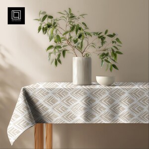 May include: A beige and white patterned tablecloth covers a wooden table. A white ceramic vase with a plant and a small white bowl are on the table. The tablecloth has a geometric design.