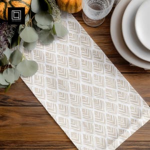 May include: A beige and white patterned table runner with a geometric design. The runner is placed on a wooden table, with pumpkins, plates, and a glass of water in the background. The pattern is a repeating series of angled lines.