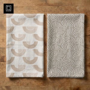 May include: Two folded kitchen towels with geometric patterns. One towel is white with tan arches, and the other is tan with white leaf-like designs. The towels are displayed on a wooden surface.