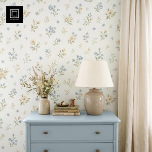May include: A light blue chest of drawers with a beige lamp, books, and a vase of flowers. The wall is covered in floral wallpaper with blue and yellow flowers. A beige curtain hangs on the right.