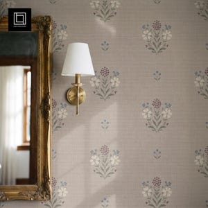 Puede incluir: Una pared cubierta con papel tapiz color topo con un patrón floral repetitivo en tonos azul, crema y verde apagados. Un espejo con marco dorado cuelga a la izquierda y una lámpara de pared con pantalla cónica blanca está fijada al papel.