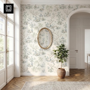 May include: An oval mirror with a thin gold frame mounted on a wall covered in off-white wallpaper featuring a repeating blue and green chinoiserie pattern of pagodas, willow trees, and birds, positioned above a potted green plant.