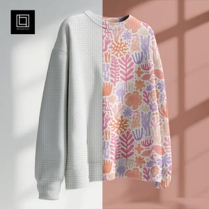 May include: A long-sleeved sweatshirt with a split design. One side is a light grey grid pattern, and the other features a colourful floral and coral reef design in pink, orange, and purple hues. The sweatshirt is displayed against a white and pink background.