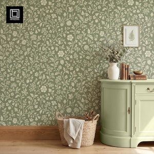 May include: A sage green floral wallpaper with a repeating pattern of cream-colored flowers and vines. A light green cabinet with a white vase, books, and a framed fern print sits against the wall. A woven basket with a white cloth is in the foreground.