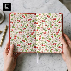 Puede incluir: Un cuaderno abierto con cubierta roja que presenta un patrón repetitivo de acuarela con cerezas de color rojo brillante, hojas verdes, flores blancas y cintas de color rosa claro sobre un fondo blanco. Incluye un marcapáginas de cinta blanca.