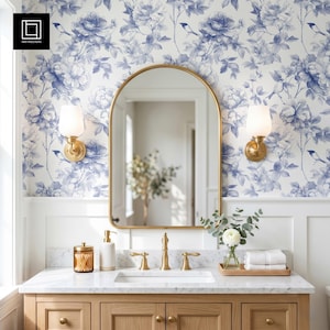 May include: A bathroom vanity featuring a light wood cabinet, a white marble countertop, and an arched mirror with a gold frame. Two wall-mounted lamps with white shades flank the mirror. The wall has blue and white floral patterned wallpaper.