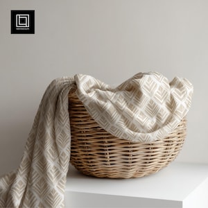 Könnte beinhalten: Ein geflochtener Weidenkorb mit einer beige-weiß gemusterten Decke. Die Decke hat ein sich wiederholendes geometrisches Design. Der Korb steht auf einer weißen Oberfläche. Die Ästhetik ist neutral und minimalistisch.