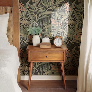 Puede incluir: Una mesita de noche de madera con un cajón y patas cónicas. La mesita de noche está junto a una cama con ropa de cama blanca y un papel tapiz con estampado floral. Un jarrón de cerámica blanco con una pequeña planta, un difusor de cerámica blanco y un reloj despertador redondo con manecillas negras están encima de la mesita de noche.