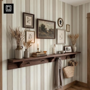 May include: A long wooden wall shelf with coat hooks featuring a collection of framed art, ceramic vases with dried grasses, a lit candle on books, a glass bottle, a gray scarf, and a woven straw tote bag hanging from the shelf hooks.