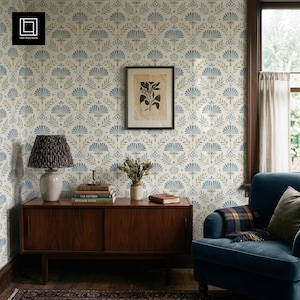Puede incluir: Un interior de habitación con papel tapiz de patrón floral azul y beige, un aparador de madera, una lámpara de mesa con pantalla plisada oscura, una lámina botánica enmarcada, un jarrón con flores secas y un sillón azul con una manta.