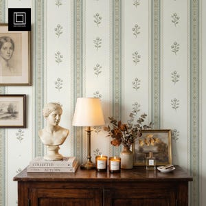 Puede incluir: Un mueble de madera oscura sostiene un busto blanco, una lámpara de latón con pantalla crema, dos libros apilados, dos velas encendidas en frascos, un arreglo floral seco en un jarrón y un cuadro de paisaje, frente a un papel tapiz de rayas.